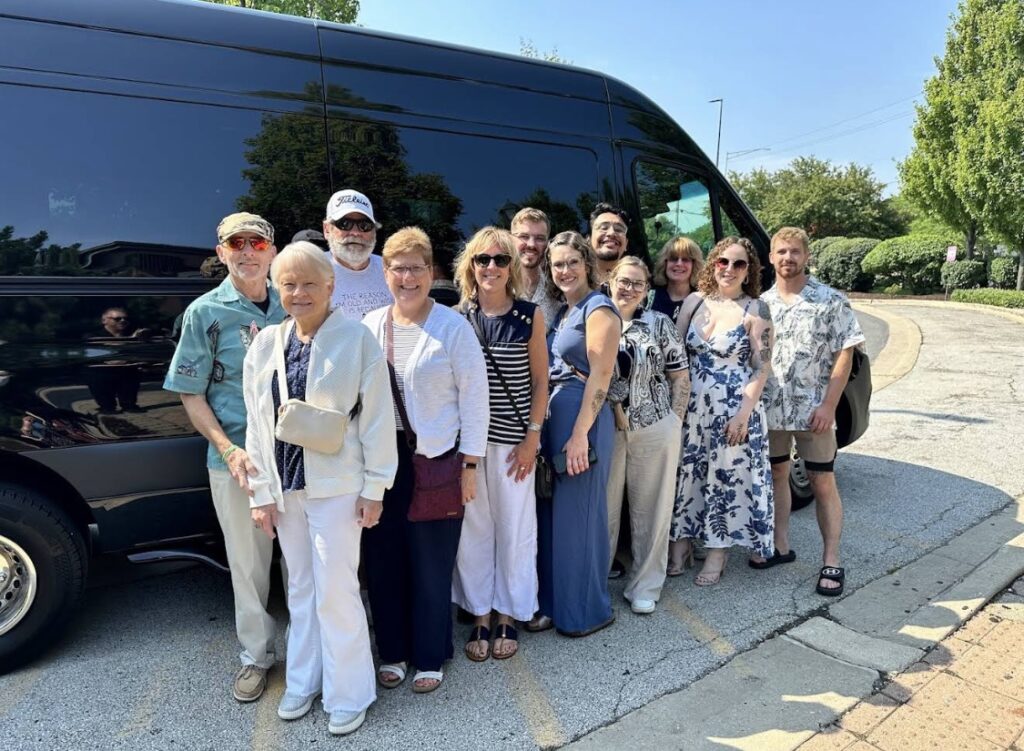 A group of twelve smiling people posing for a photo in front of a sleek, black executive passenger van from Rockford Limo Company. The group is standing on a sunny day in a paved parking area with green trees in the background, showcasing the company's ability to handle large group transportation and special events.
