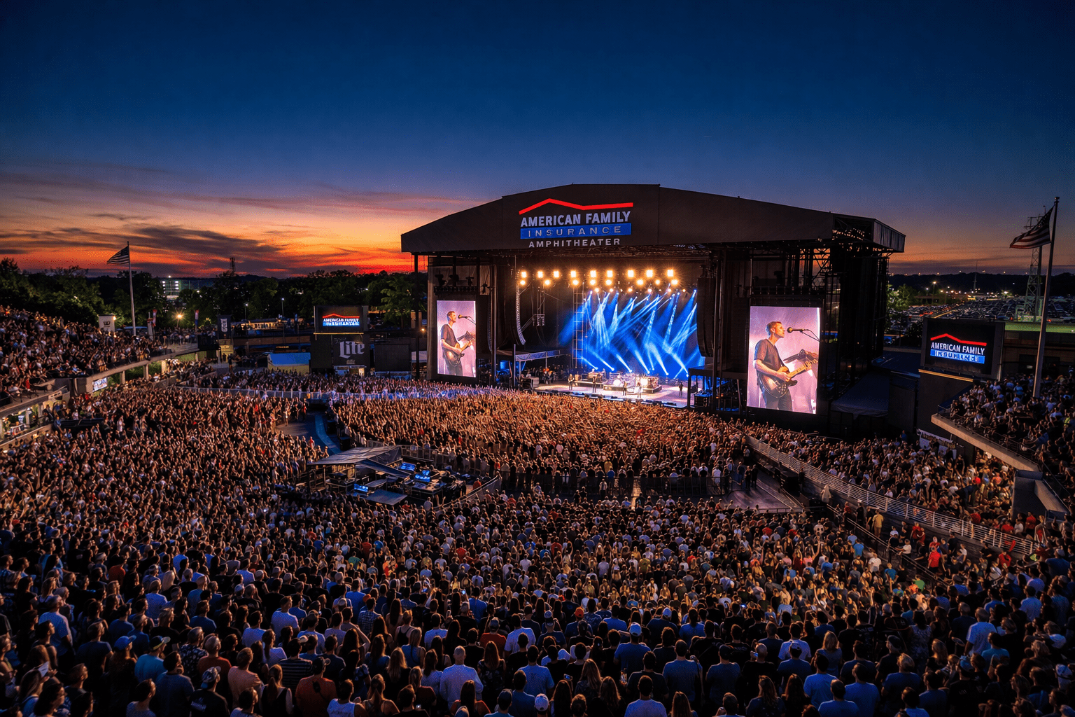 Large crowd at a concert inside the American Family Insurance Amphitheater at dusk, with a brightly lit stage and massive video screens.